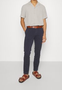 Camicia grigia chiara a maniche corte, pantaloni blu navy, cintura marrone e sandali marroni con design a fascia e suole nere.