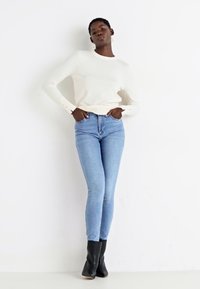 Cream fitted long-sleeve top paired with light blue skinny jeans and black ankle boots. Model stands against a plain white background.