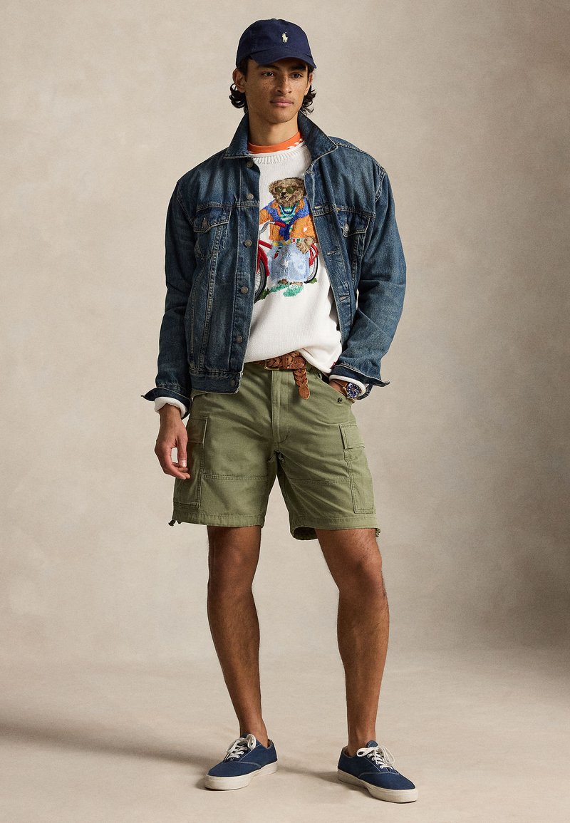 Jeune homme portant une veste en denim, un pull avec un ours à vélo, un short cargo olive, une casquette marine et des baskets marines, debout devant un fond uni.