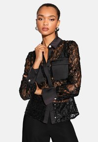 Crna čipkasta bluzica izrađena od prozirnog materijala, s satenskim ovratnikom i manžetama. Ima kopčanje na dugmad i džepove, ukrašena dekorativnim dugmadi.