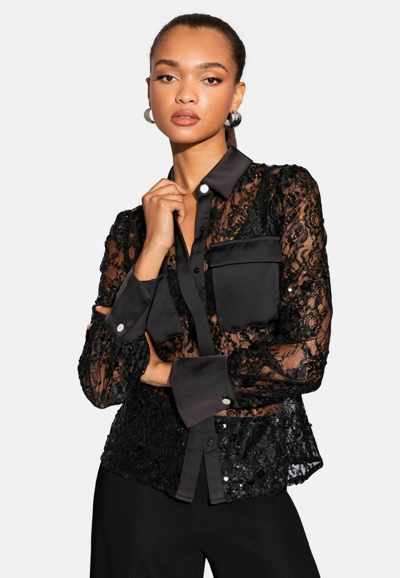Crna čipkasta bluzica izrađena od prozirnog materijala, s satenskim ovratnikom i manžetama. Ima kopčanje na dugmad i džepove, ukrašena dekorativnim dugmadi.