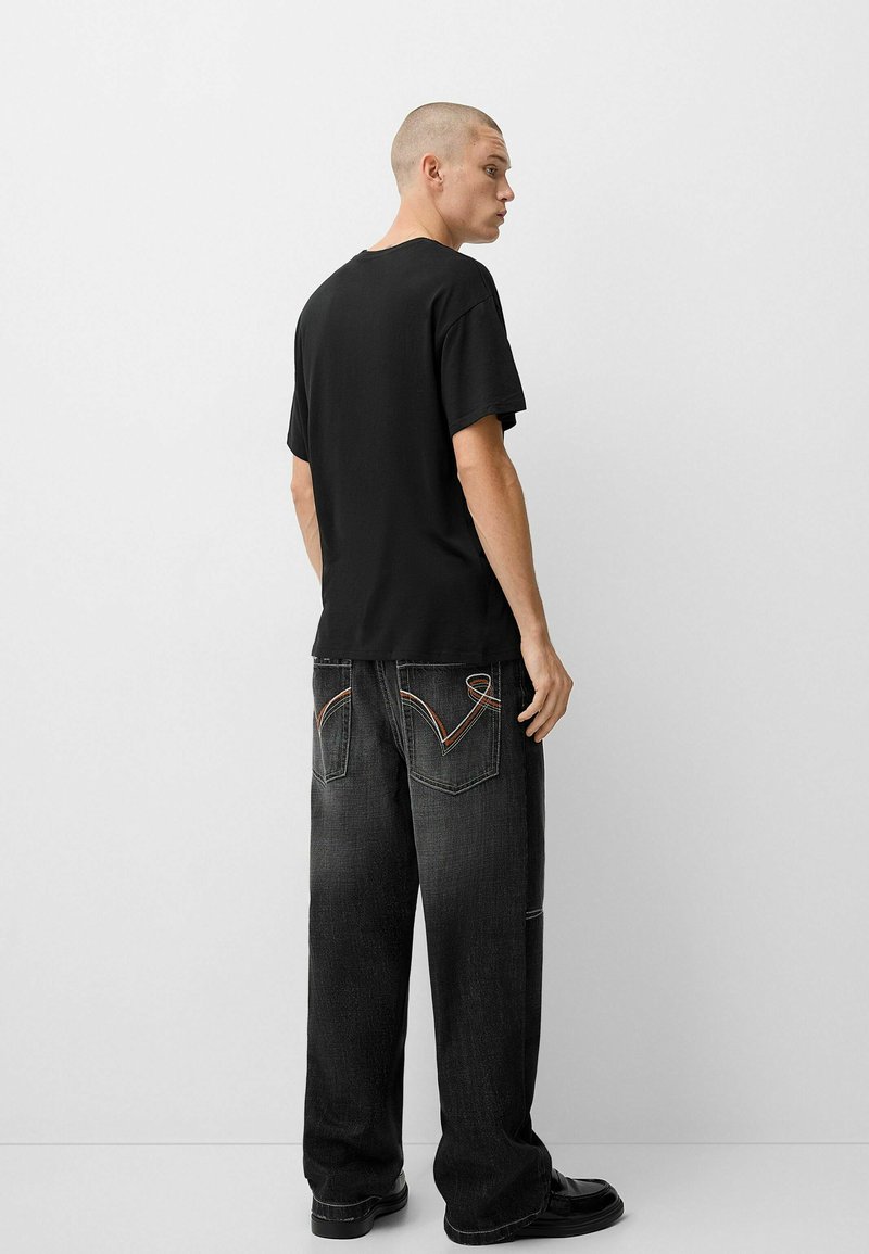 Jeune homme avec la tête rasée portant un t-shirt noir simple, un jean noir ample avec des designs de poches cousus et des chaussures noires sur fond blanc.