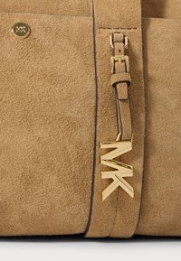 Tan zomšos rankinė su auksiniu apdailos akcentu ir "MK" logotipu, odiniu dirželiu su auksiniu užsegimu ir tekstūruota paviršiumi.