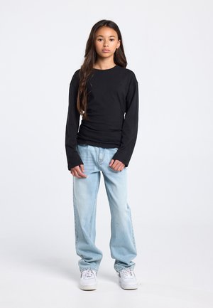 Chemise noire à manches longues, coupe ajustée, associée à un jean bleu clair ample et des baskets blanches, posant devant un fond uni.