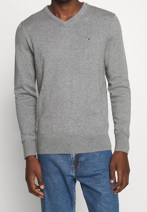 Hombre con suéter gris de cuello en V con un pequeño logo en el pecho y jeans azules, de pie contra un fondo liso.