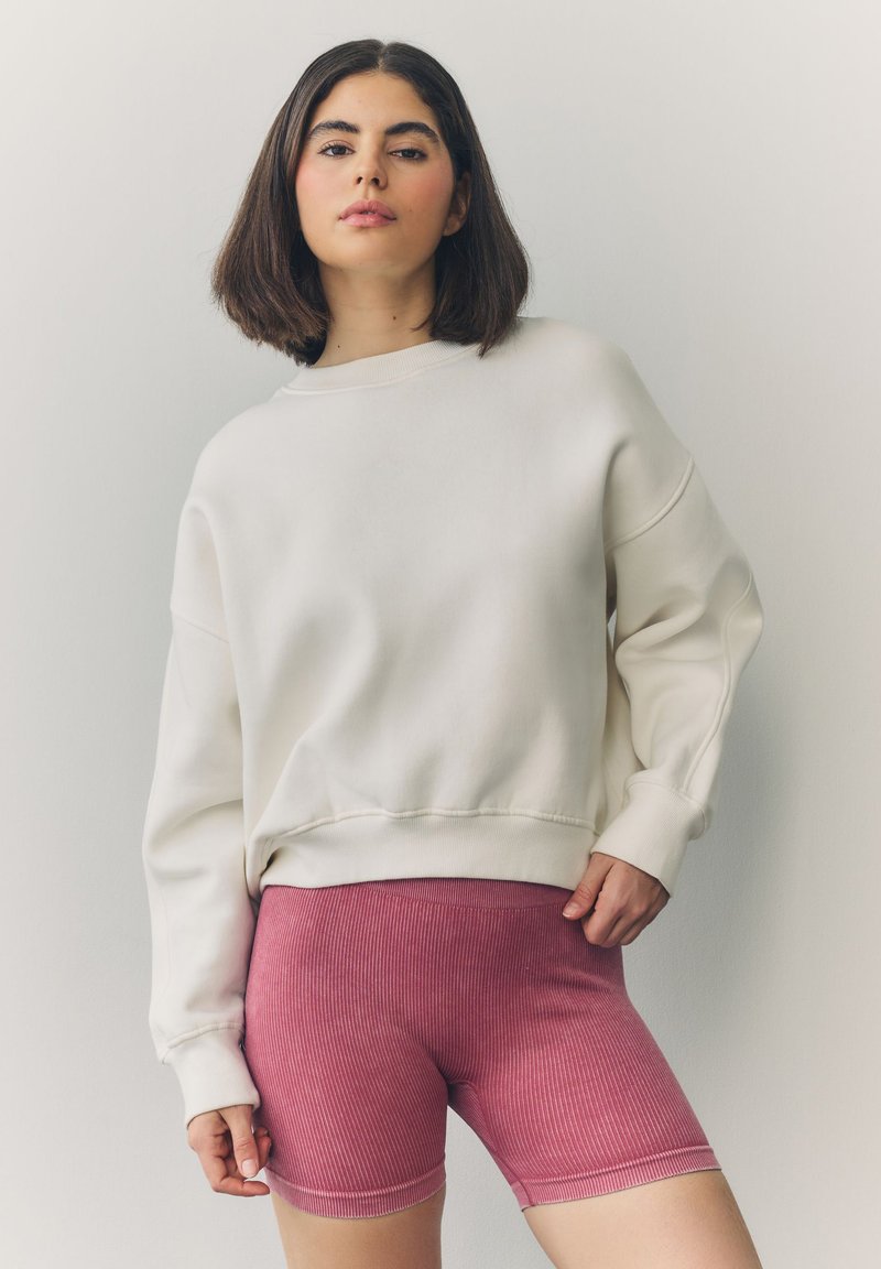 Sudadera blanca con hombros caídos, ajuste relajado, puños acanalados, combinada con pantalones cortos tipo ciclista de punto acanalado de color rosa claro. Diseño simple y casual.