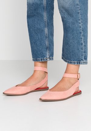 Fødder iført lyserøde flade sko med spidse tæer og ankelstropper, kombineret med blå cropped denim-jeans mod en ensfarvet baggrund.