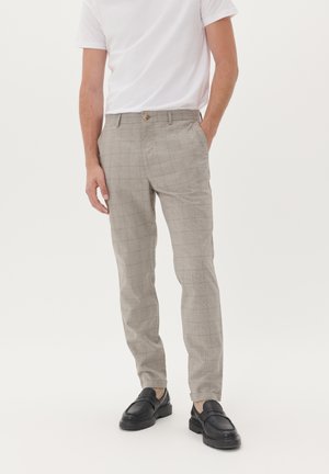 Pantalones de cuadros en un tono beige claro con cierre de botón, bolsillos laterales y un ajuste entallado, combinados con zapatos negros de slip-on y una camiseta blanca lisa.