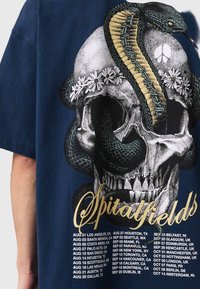 Granatowy T-shirt z ilustracją czaszki ozdobionej stokrotkami i wężem, uzupełniony tekstem wymieniającym daty trasy koncertowej w kolorach złotym i białym.