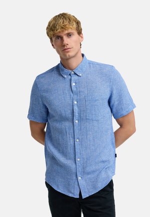 Ung mand med kort blondt hår iført en blå kortærmet skjorte med lomme, står med hænderne bag ryggen.