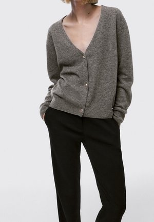 Frau trägt eine graue V-Ausschnitt Strickjacke mit Knöpfen und schwarze Hosen, steht mit Händen in den Taschen vor einem einfarbigen hellen Hintergrund.