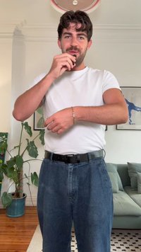 Λευκό κοντομάνικο πουκάμισο, μαύρη ζώνη, φαρδιά μπλε τζιν παντελόνια, χρυσό βραχιόλι στον καρπό, στέκεται σε εσωτερικό χώρο με φυτά και έναν καναπέ στο παρασκήνιο.