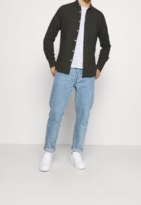 Camicia a maniche lunghe grigio scuro con bottoni sopra una T-shirt grigio chiaro, abbinata a jeans azzurri risvoltati e sneaker bianche.