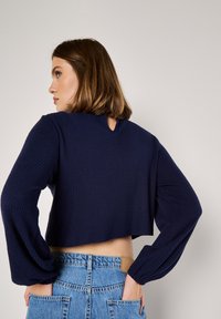 Marineblauer, cropped Pullover mit gerippter Textur und Rundhalsausschnitt. Er hat lange Pompom-Ärmel und ein Schlüssellochdetail auf der Rückseite.
