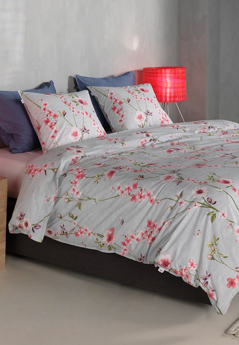 Květinový povlečení s jemně šedým pozadím a vzorem růžových a zelených květin. Dvě odpovídající povlaky na polštáře a dřevěný noční stolek.