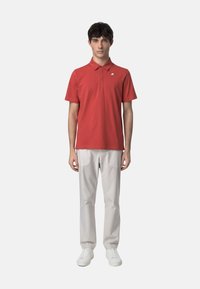Polo rouge avec col, manches courtes et petit logo sur la poitrine ; associé à un pantalon gris clair et des baskets blanches.