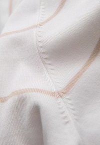 Valkoinen kangas, jossa on hienovaraisia beigejä raitoja, pörröisellä kudonnalla ja näkyvillä ompelutiedoilla. Pehmeä ja kevyt materiaali.