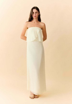 Femme aux longs cheveux foncés portant une robe maxi crème sans bretelles et des sandales beiges, debout devant un fond beige uni.