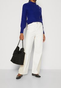 Pull en tricot bleu roi avec un col montant et des détails à boutons, associé à un pantalon ample blanc cassé et un sac à main noir texturé.