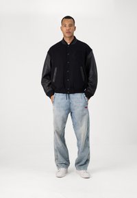 Blouson noir avec des manches en cuir contrastantes, arborant un logo texturé. Associé à un jean en denim bleu clair et des baskets blanches.