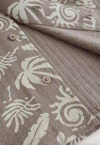 Hellbrauner Stoff mit weißen tropischen und abstrakten Mustern, zeigt vernähte Kanten und eine strukturierte Oberfläche.