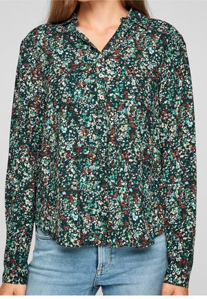 Nainen, jolla on vihreä kukallinen pitkähihainen nappipaita yhdistettynä vaaleansinisiin farkkuihin, jotka ovat leikattu keskikropan kohdalta.