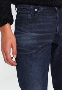 Tmavě modré džíny s úzkým střihem, vybavené předními a zadními kapsami, kovovým knoflíkem a jemnými detaily v oblasti vyblednutí. Texturovaná látka.