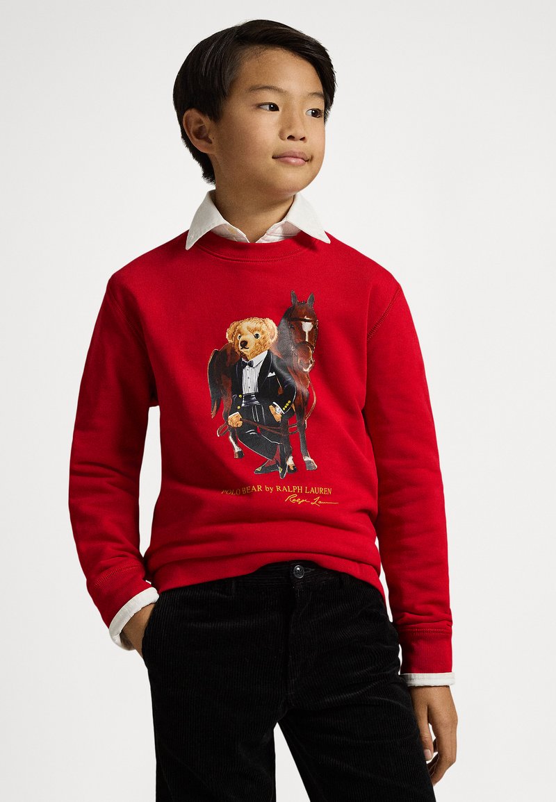 Sweatshirt vermelho com estampa gráfica de um urso de smoking ao lado de um cavalo. Texto e detalhes em amarelo e preto. Calças de bombazine pretas.