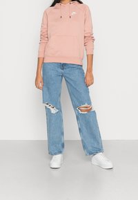 Ljusrosa hoodie med framficka och vitt logotyp, kombinerad med slitna blå jeans och vita sneakers, stående på en enkel yta.