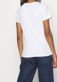 Persona de piel oscura y cabello trenzado lleva una camiseta blanca de manga corta sin estampado y unos vaqueros oscuros de denim sueltos, mostrada desde atrás.