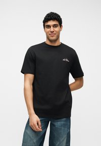 Νεαρός με σγουρά μαλλιά που φοράει μαύρο κοντομάνικο T-shirt με το λογότυπο «Le Deux» και μπλε τζιν με σχέδιο patchwork, στέκεται μπροστά σε λευκό φόντο.