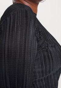 Top plissettato nero con una texture liscia, scollatura rotonda e dettagli delle maniche arricciati, caratterizzato da una silhouette aderente che mette in risalto le pieghe.