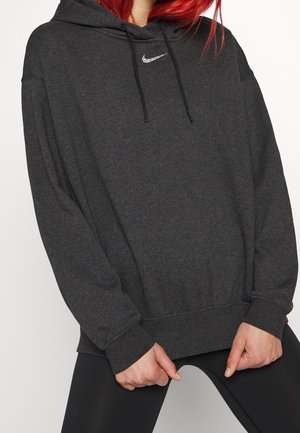 Personne aux cheveux roux portant un sweat à capuche Nike gris foncé et des leggings noirs, tenant l'ourlet du sweat à capuche des deux mains devant.