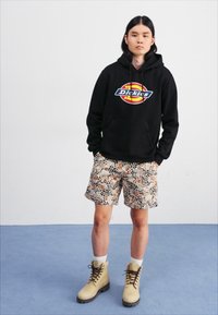 Black hoodie with colorful "Dickies" logo, patterned tan shorts with animal motifs, and beige boots with laces, set against a neutral background.