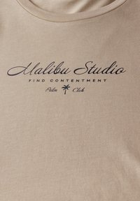 T-shirt beige clair en coton doux avec un col rond, arborant un texte imprimé noir : "Malibu Studio Find Contentment Palm Club."