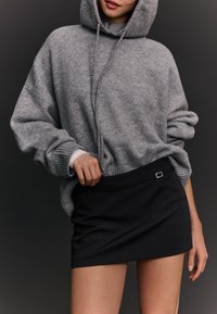 Donna che indossa un ampio maglione grigio con cappuccio e una mini gonna nera, che sistema la cintura della gonna con la mano su sfondo scuro.