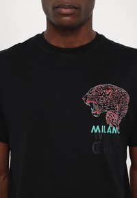 Crna pamučna majica s grafikom leoparda u crvenoj i ružičastoj boji, s natpisom "MILANO" otisnutim u tirkiznoj boji i "WILD ONES" u crnoj boji ispod.