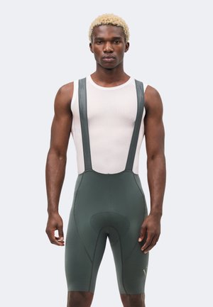 Μυώδης άνδρας με κοντά ξανθά μαλλιά, που φοράει άσπρη διχτυωτή μπλούζα χωρίς μανίκια και σκούρα πράσινα ποδηλατικά σορτς με τιράντες.