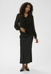 Zwarte ruffle blouse met lange mouwen, gecombineerd met een strakke zwarte satijnen maxi rok en zwarte puntige hakken; zachte textuur en elegante uitstraling.