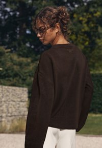 Ruskea neulottu villapaita pyöreällä pääntiellä, lasketuilla olkapäillä ja resorireunuksella. Malli käyttää vaaleita housuja ja aurinkolaseja.