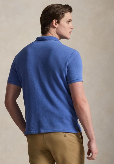 Νέος άνδρας που φοράει μπλε κοντομάνικο πόλο πουκάμισο και μπεζ παντελόνι, στέκεται πλαγίως με τα χέρια κοντά στις τσέπες, σε απλό φόντο.