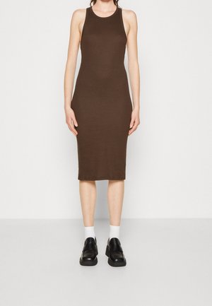 Kobieta ubrana w brązową sukienkę bez rękawów, sięgającą do kolan, białe skarpetki i czarne mokasyny, stojąca na jednolitym białym tle.