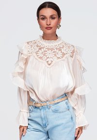 Femme portant un chemisier blanc en dentelle à col haut avec des volants et un jean taille haute bleu clair avec une ceinture beige, posant devant un fond uni.