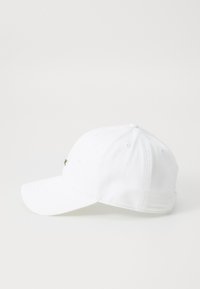 Hvit baseballcap med buet skygge, med en liten grønn brodert logo på fronten. Laget av glatt stoff med sydde paneler.