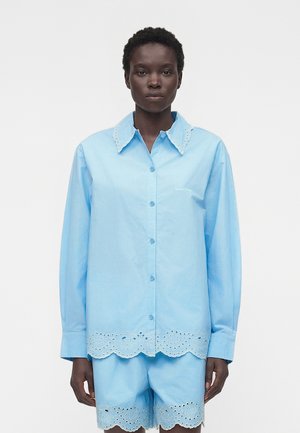 Osoba nosi svetlomodrú košeľu s dlhým rukávom na gombíky a zodpovedajúce šortky s vlnitými vyšívanými okrajmi na golieri, leme a nohaviciach.