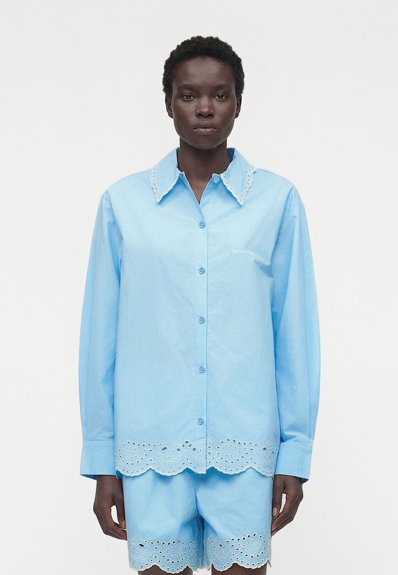 Άτομο που φοράει ανοιχτομπλε πουκάμισο με μακριά μανίκια και κουμπιά καθώς και ταιριαστό σορτς με κυματιστές λεπτομέρειες με κεντήματα στην γιακά, το τελείωμα και τα πόδια.