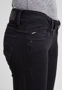 Black denim jeans featuring a fitted shape, zip fly, and small "RAW" logo detail near the pocket. Textured fabric with a smooth finish.