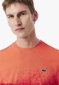 Orange t-shirt med en gradientdesign, texturerad tyg och en liten grön och röd logotyp på vänster bröstområde.