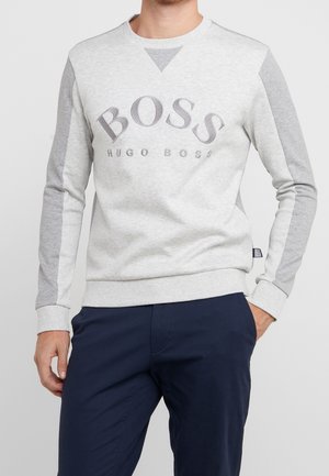 Mężczyzna ubrany w jasno- i ciemnoszary długi rękaw bluzy Hugo Boss z granatowymi spodniami, ręka w kieszeni, neutralne tło.