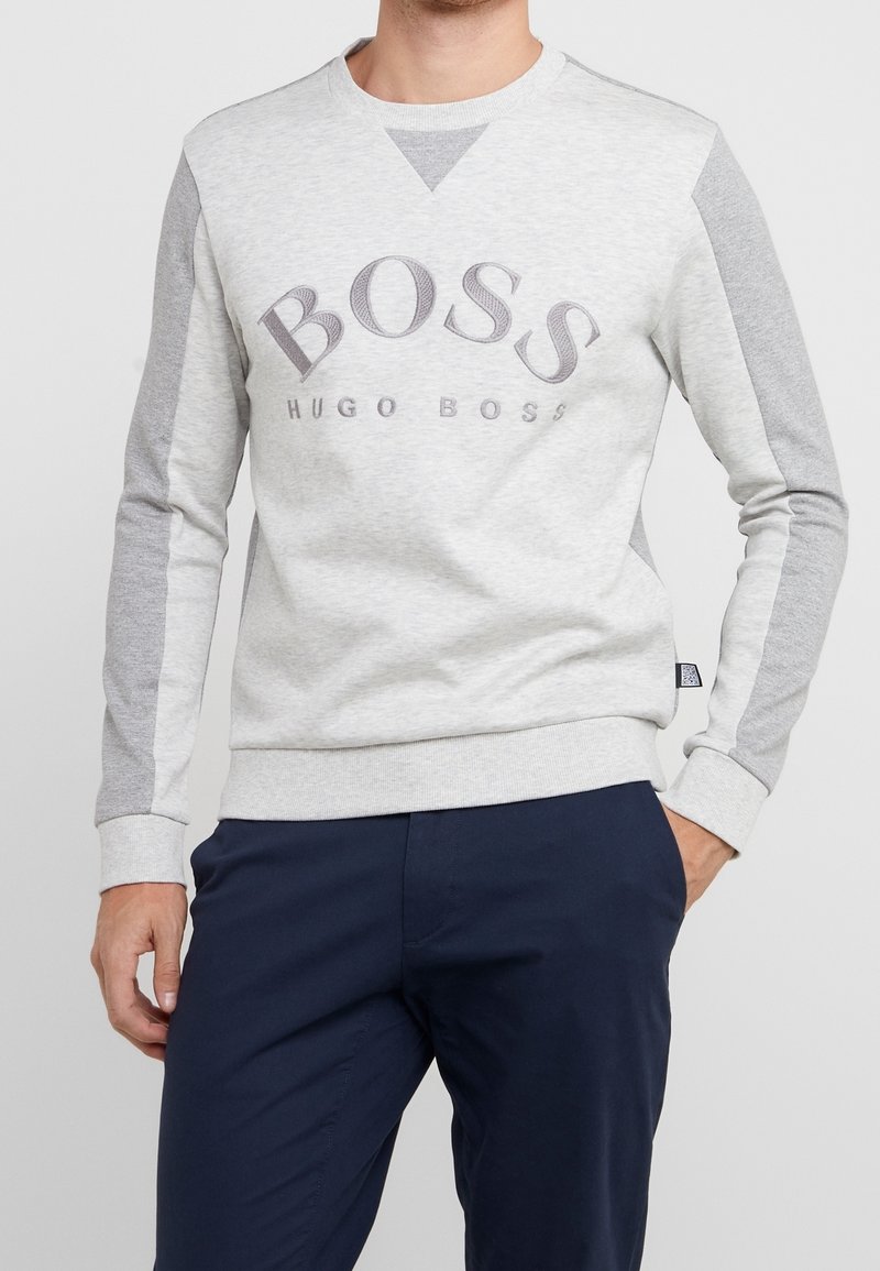 Homme portant un sweat-shirt Hugo Boss à manches longues gris clair et gris foncé avec un pantalon bleu marine, main dans la poche, fond neutre.
